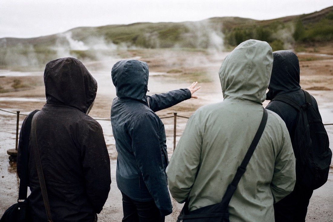 A guide pointing out the rope line to a small group at the Geysir geothermal field, seen from behind in the steam