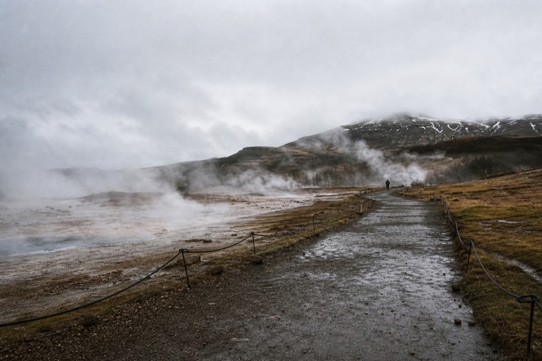 Wet gravel path with wooden posts and rope lines winding through the steaming Geysir geothermal field in drizzle