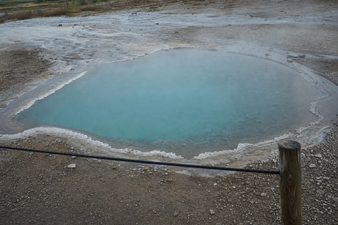 Silica-blue hot spring pool at the Geysir geothermal area behind a low rope barrier, steam drifting off the surface