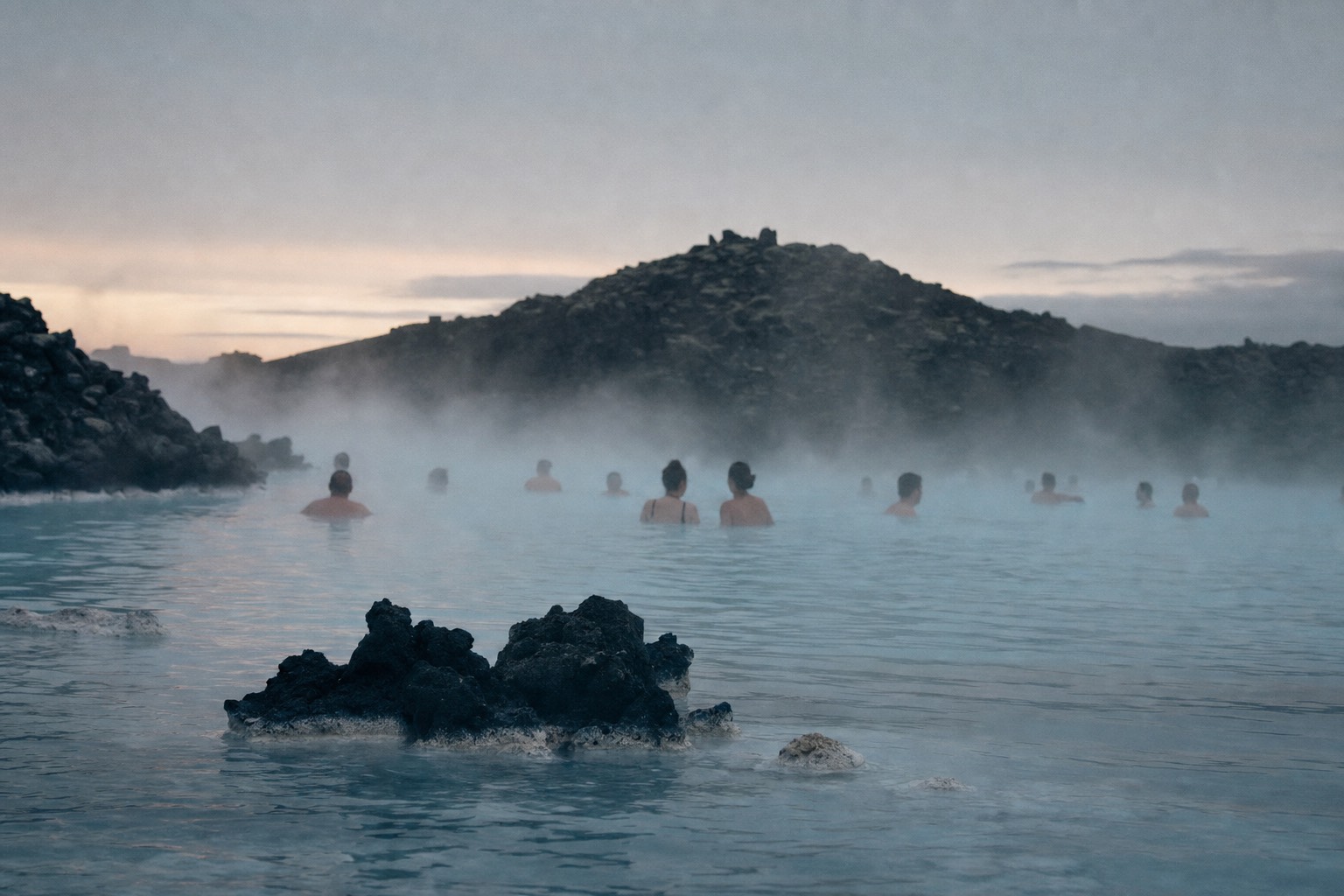 Steam drifting over the milky blue water of the Blue Lagoon at dusk, dark lava field behind