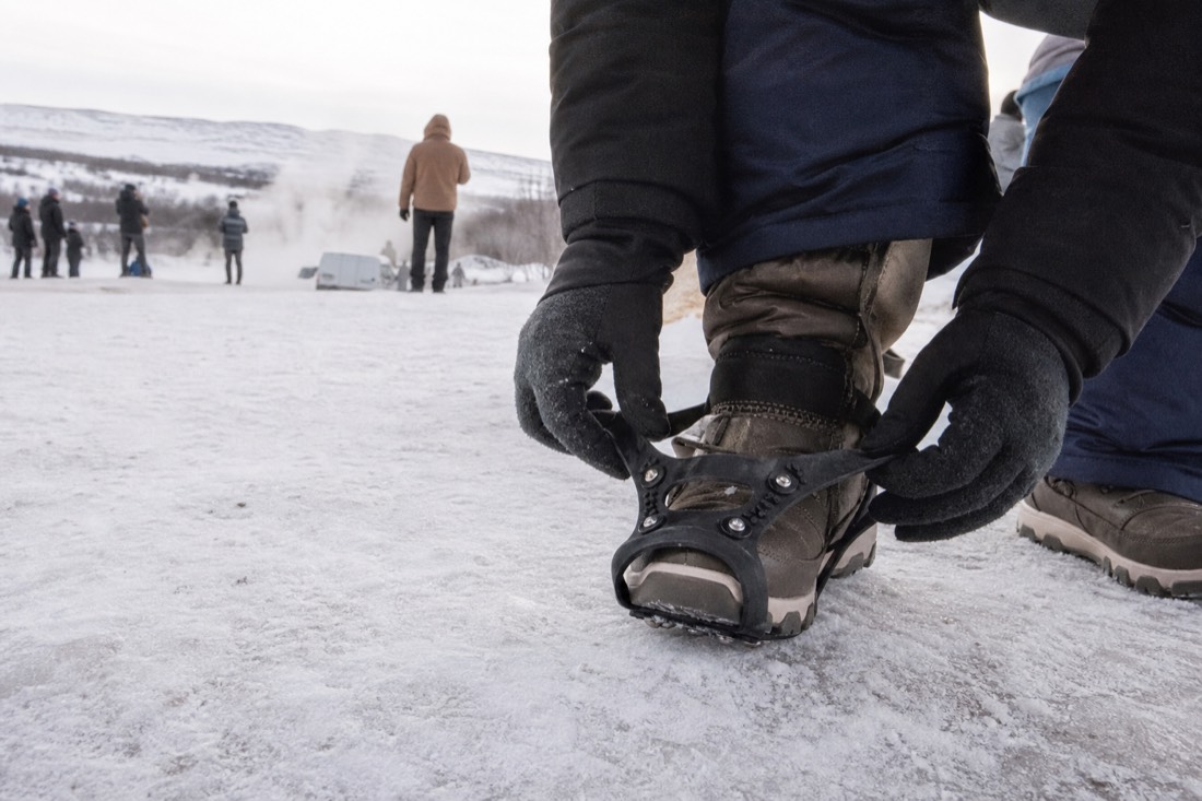 Strap-on ice grips being fitted over winter boots on packed snow at the Geysir field