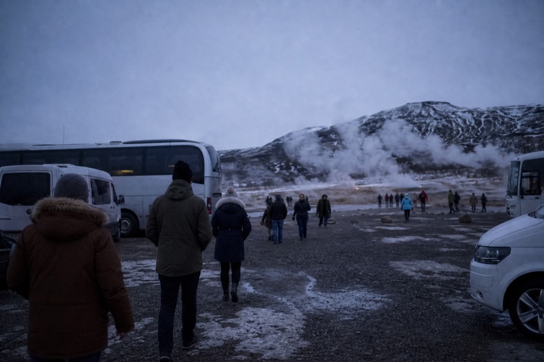 Coaches and small tour vans in the gravel lot at Geysir with steam drifting behind at winter dusk