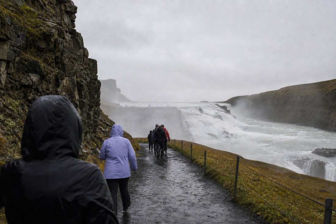 Travelers in rain jackets walking the lower path beside the two-tier cascade of Gullfoss in drizzle