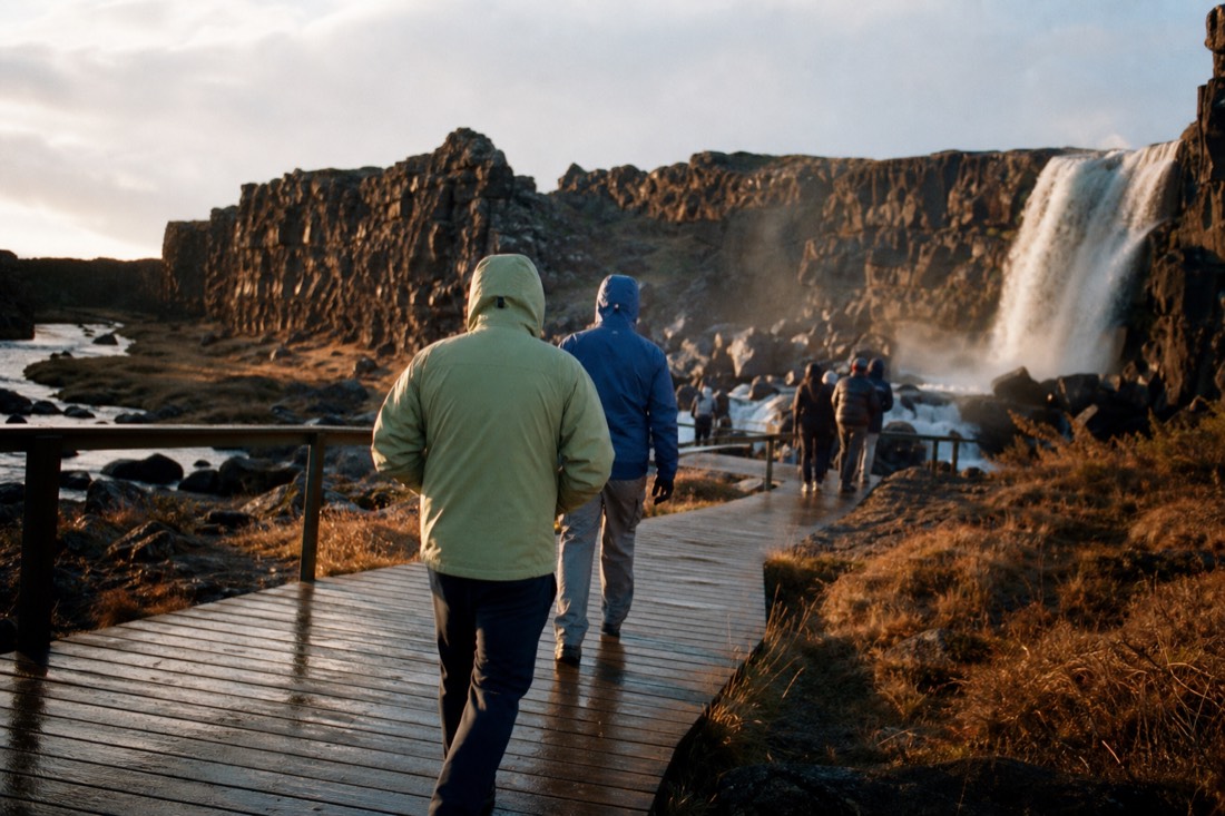 Hikers in shell jackets on the boardwalk path toward Öxarárfoss in low evening light